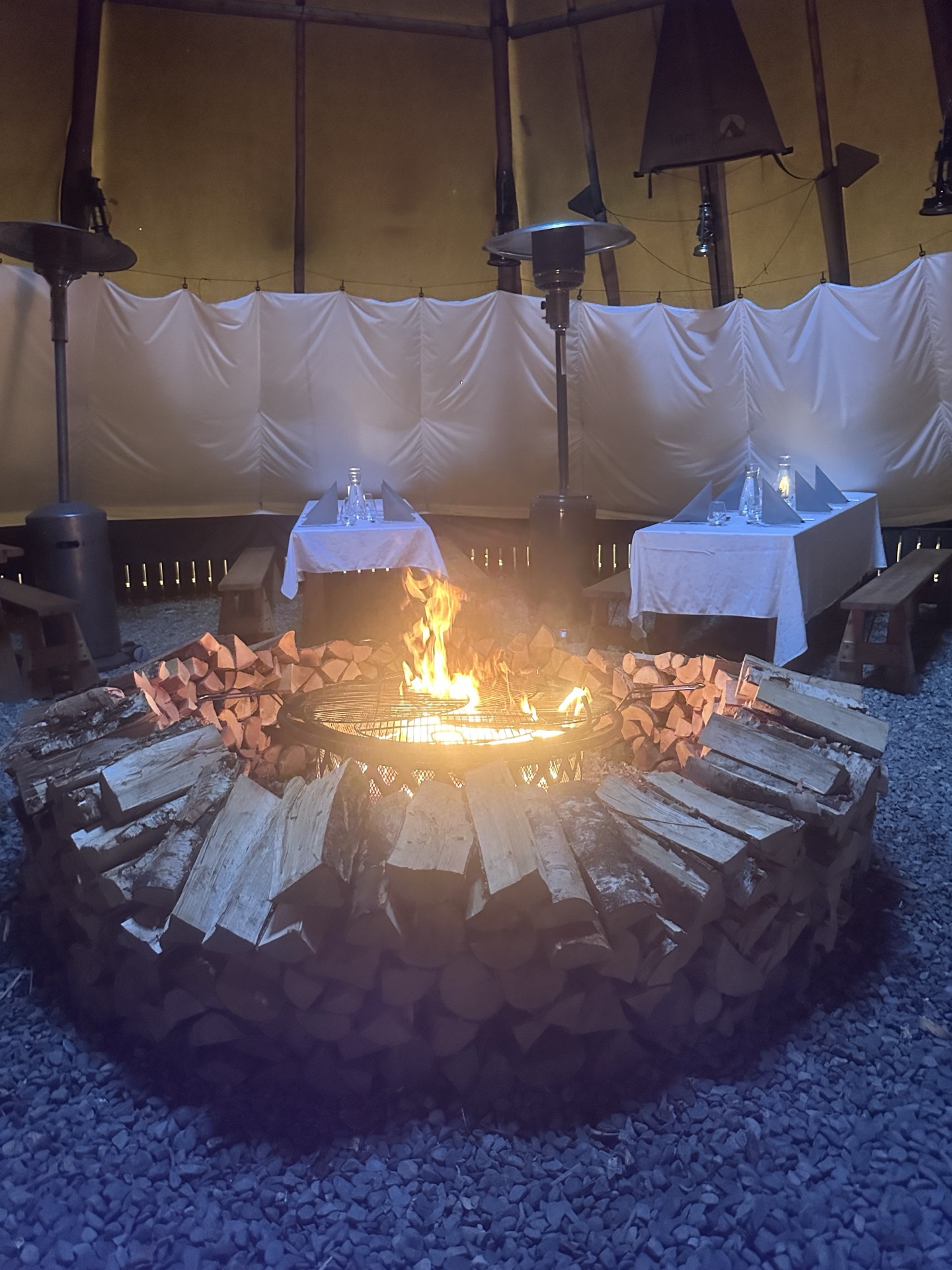 A lit fire pit ringed with stacked logs inside a tipi laid for dinner.